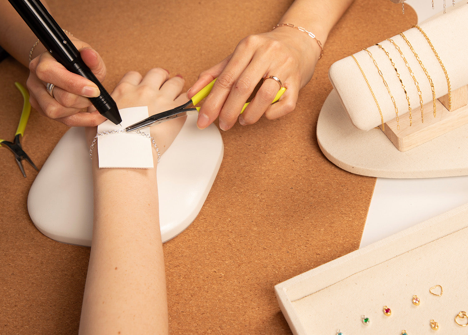 a jewellery designer fitting a permanent bracelet to a customer, using a precision welder. On display are a selection of sterling silver and 14ct gold filled chains and charms for the customer to choose from
