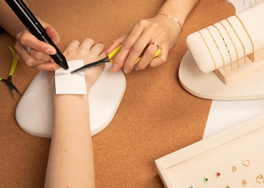 a jewellery designer fitting a permanent bracelet to a customer, using a precision welder. On display are a selection of sterling silver and 14ct gold filled chains and charms for the customer to choose from